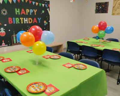 Image showing a colorfully decorated room with a Happy Birthday banner on the wall, tables are decorated with balloons, and colorful plates and napkins.
